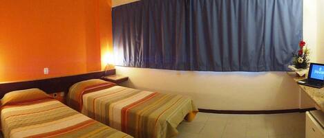 Habitación de hotel con dos camas, un escritorio con una laptop, una ventana con cortinas y una pequeña planta en el escritorio.