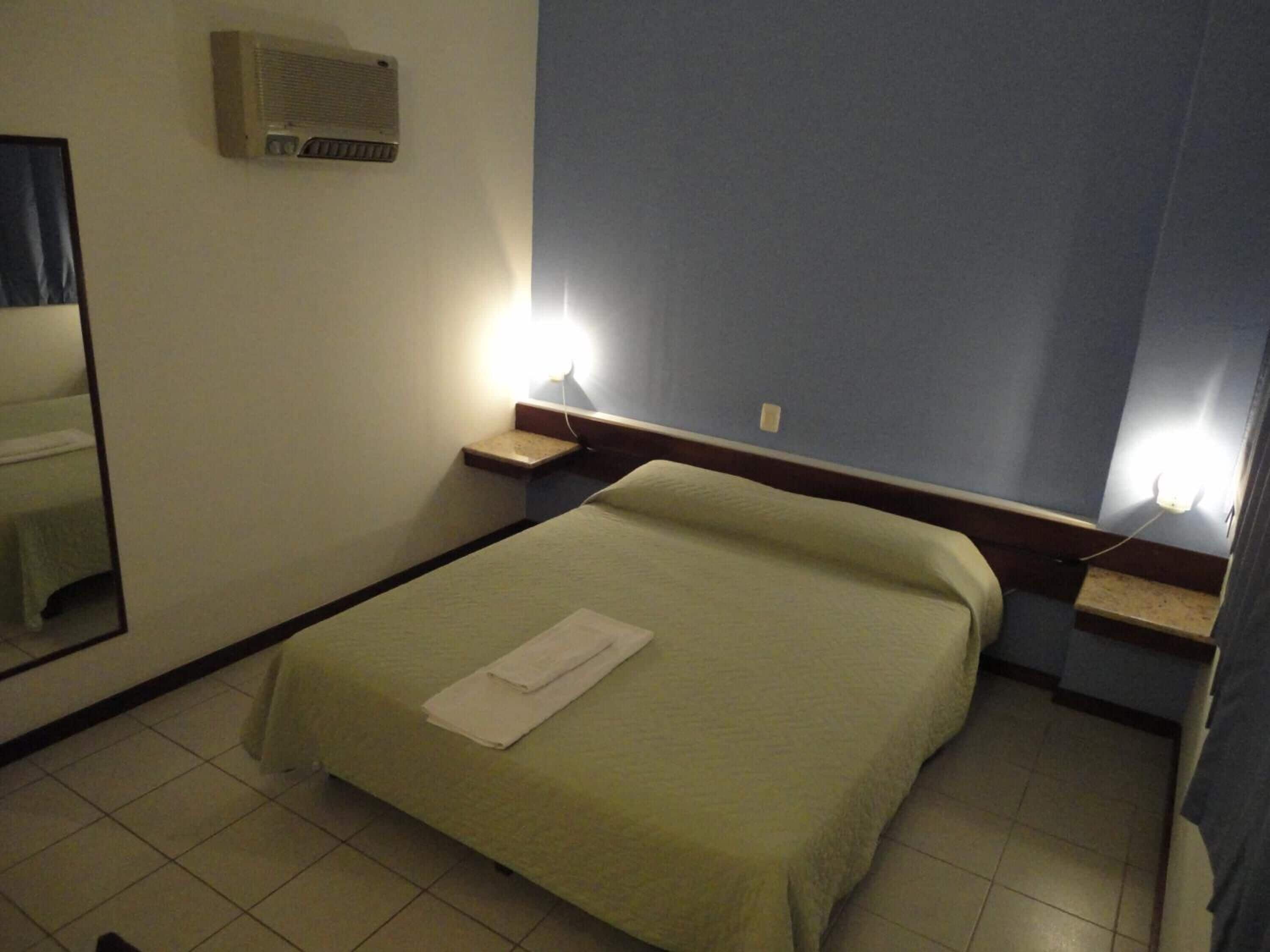 A hotel room with a bed, a nightstand, a mirror, and an air conditioning unit on the wall.