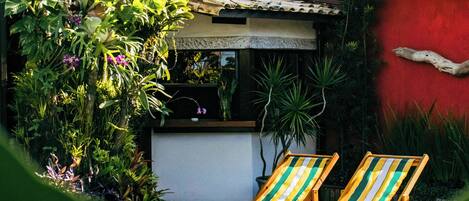 A pool area with two lounge chairs, surrounded by lush greenery and a red building with a tiled roof.