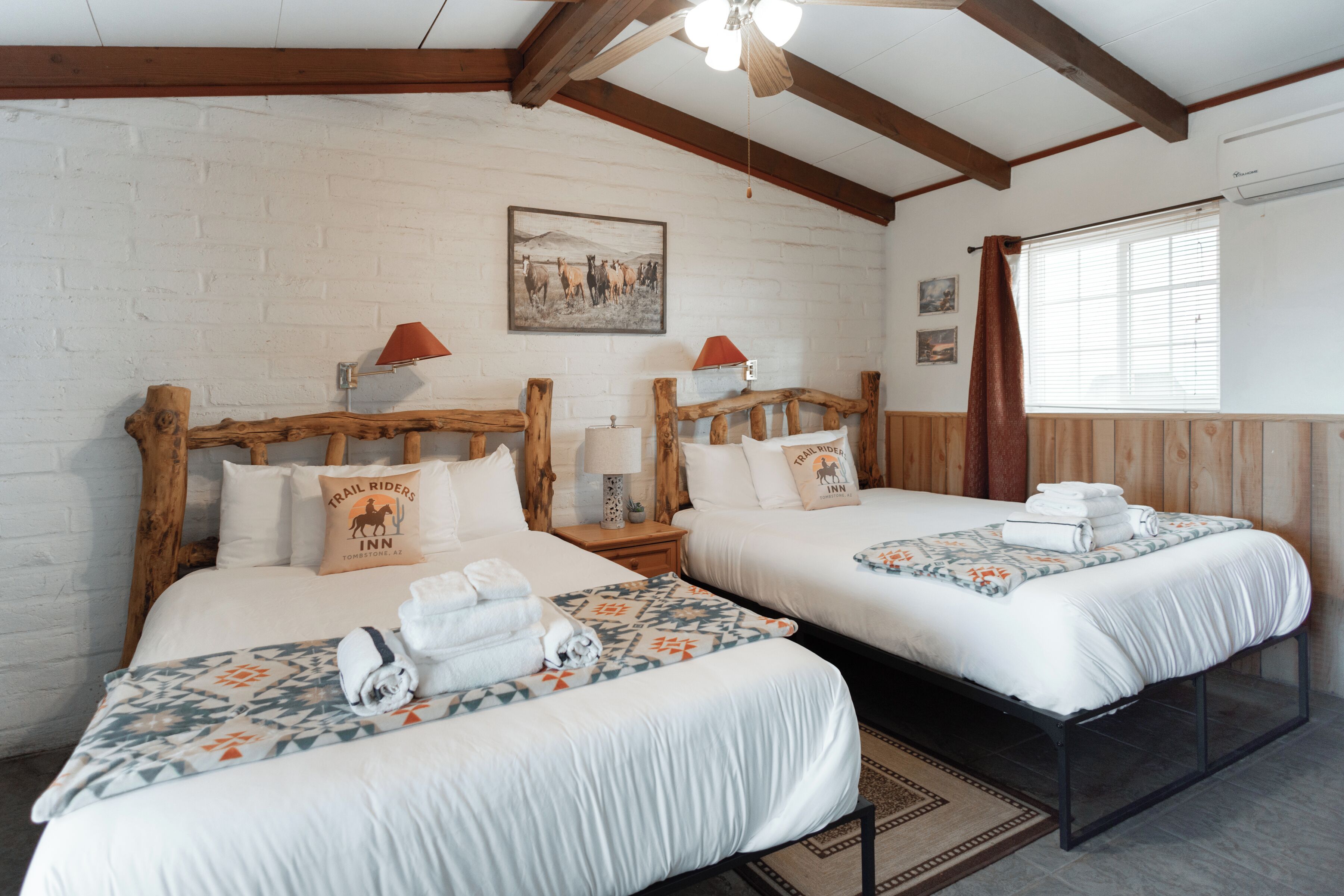 A hotel room with two beds, wooden headboards, white linens, a mounted picture, and a window with blinds.