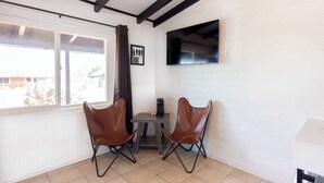 A room with two brown leather chairs, a small table with a coffee maker, a TV mounted on the wall, and a window with a view of buildings outside.