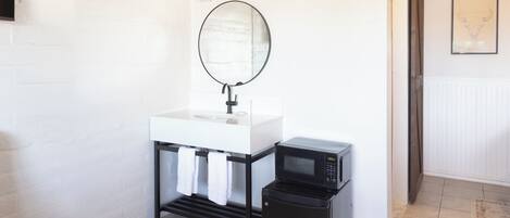 Une petite salle de bain avec un lavabo blanc, un miroir, un micro-ondes noir et un petit réfrigérateur noir.