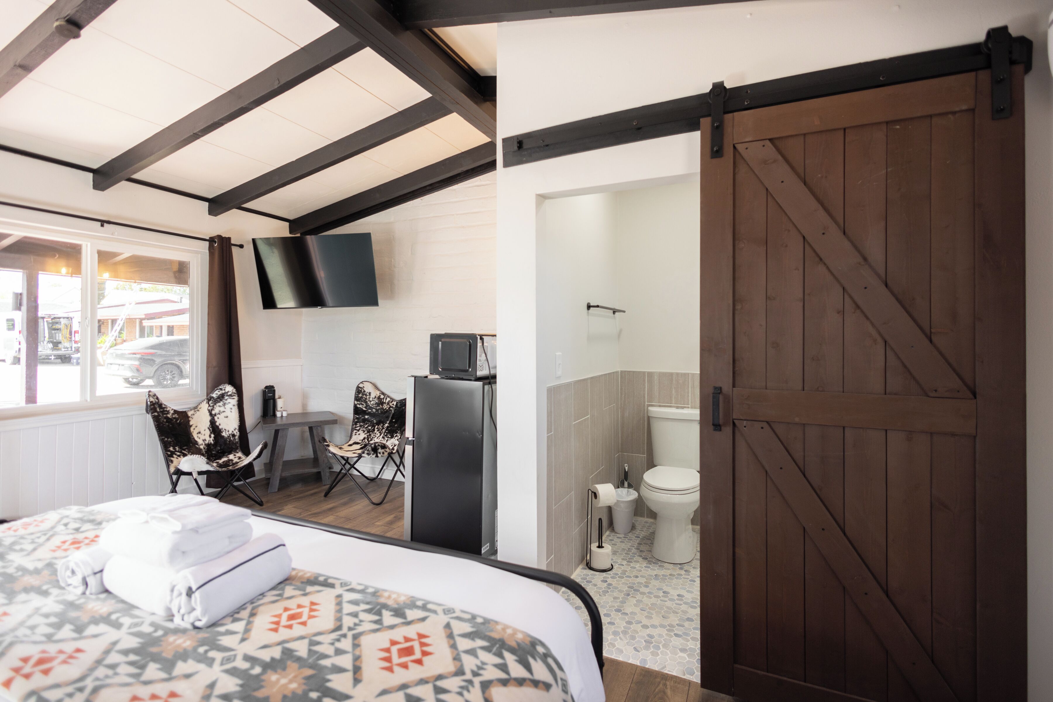 A hotel room with a bed, a desk, a chair, a TV, and a bathroom with a wooden door.
