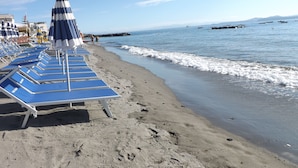 A beach with rows of blue lounge chairs and striped umbrellas, a sandy shore, and calm sea with boats in the distance. - Villa Panoramica (Ischia)