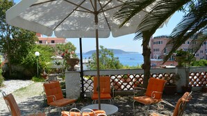 An outdoor seating area with orange chairs and a white umbrella, overlooking a coastal view with buildings and a body of water. - Villa Panoramica (Ischia)