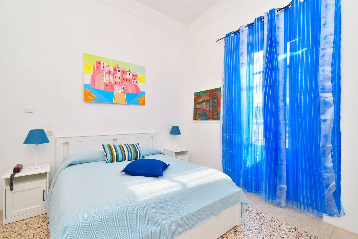 Ein Schlafzimmer mit einem Bett, zwei Nachttischen, einem Beistelltisch, einem blauen Vorhang und zwei Bildern an der Wand.