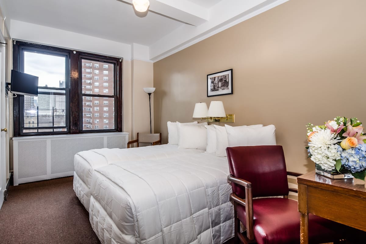 A hotel room with a large bed, a desk with a chair, a window with a view of buildings, a floor lamp, and a framed picture on the wall.