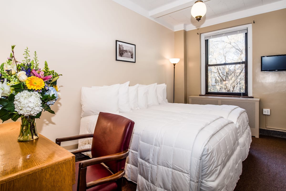 A hotel room with a bed, a wooden table with a vase of flowers, a chair, a standing lamp, and a window with a view of trees.