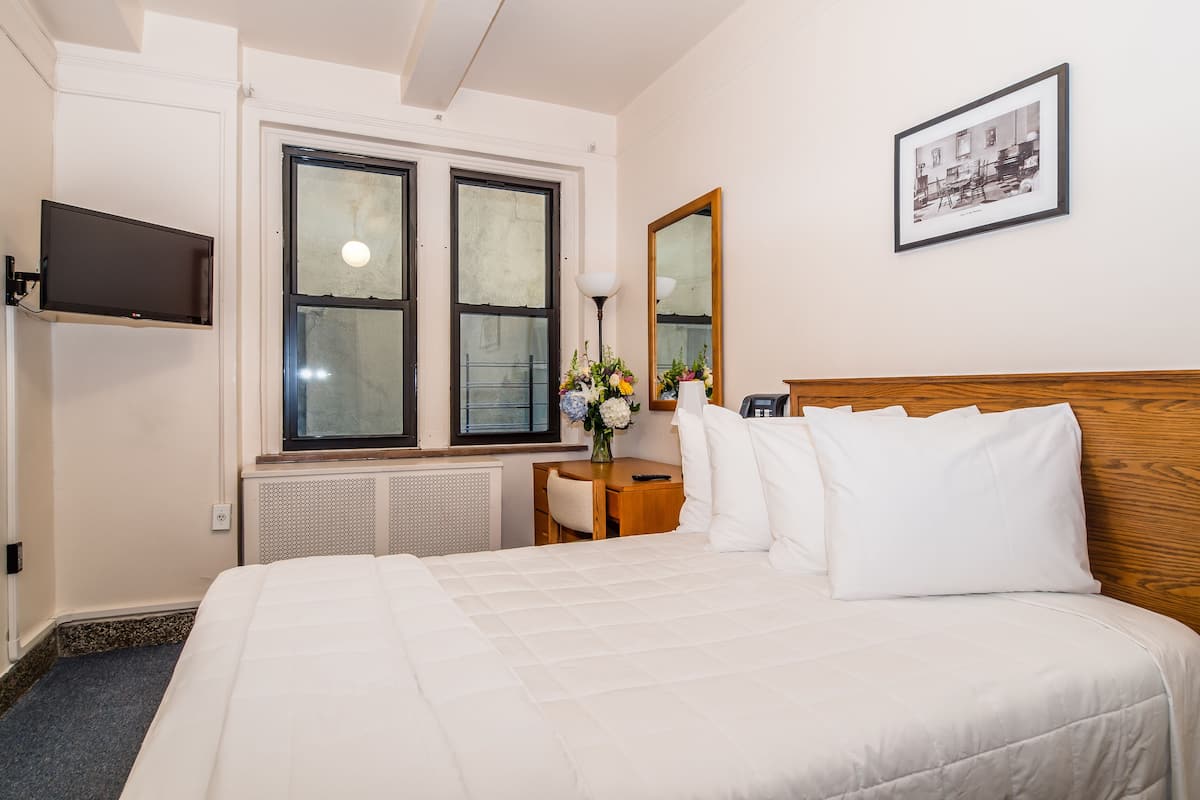 A hotel room with a large bed, a flat-screen TV mounted on the wall, a wooden headboard, a nightstand with a lamp, and a framed picture on the wall.