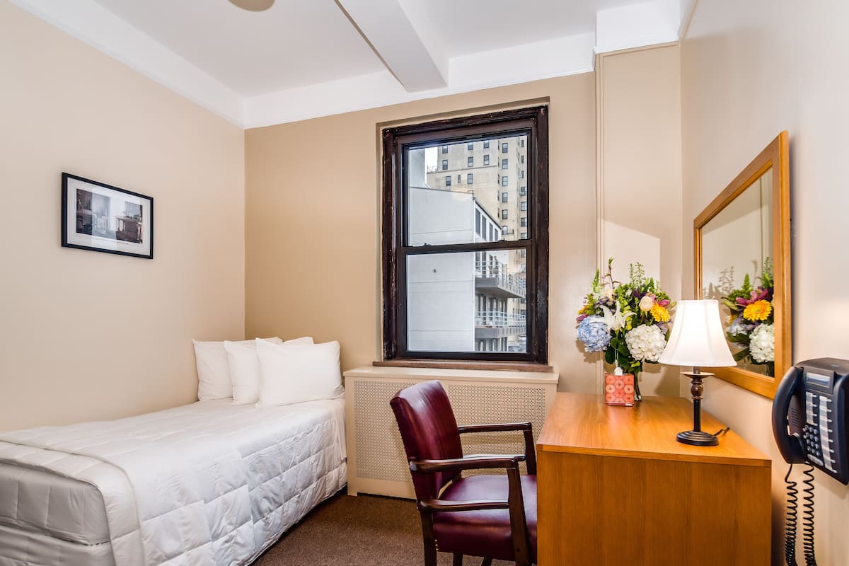A hotel room with a bed, a desk with a telephone, a chair, a window with a view of buildings, and a framed picture on the wall.