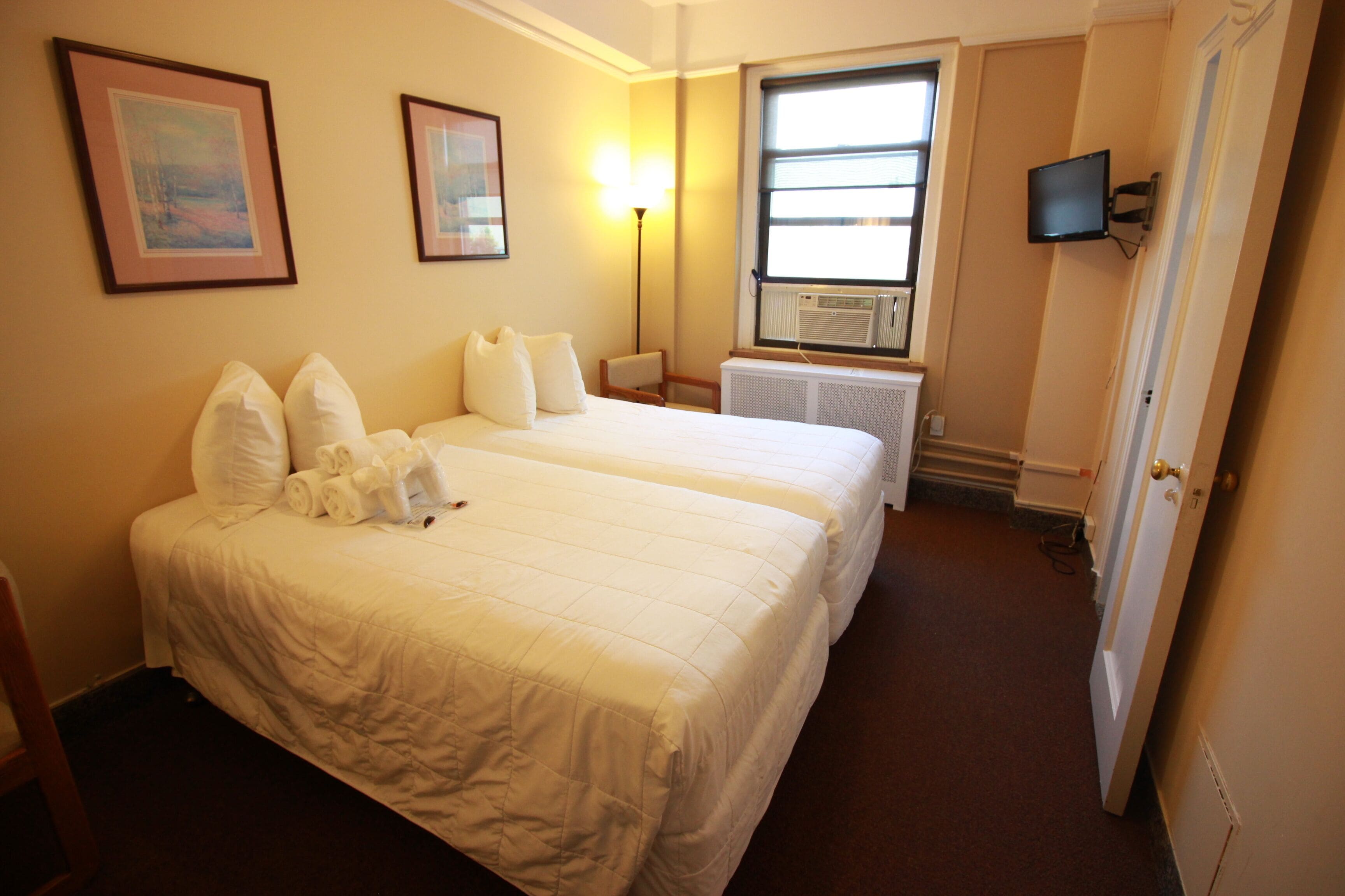 A hotel room with two beds, a TV mounted on the wall, a window with a view, and a radiator.