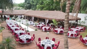 An outdoor dining area with round tables and red chairs, surrounded by palm trees and covered by thatched umbrellas. - Lanville Athenee (Foz do Iguacu)