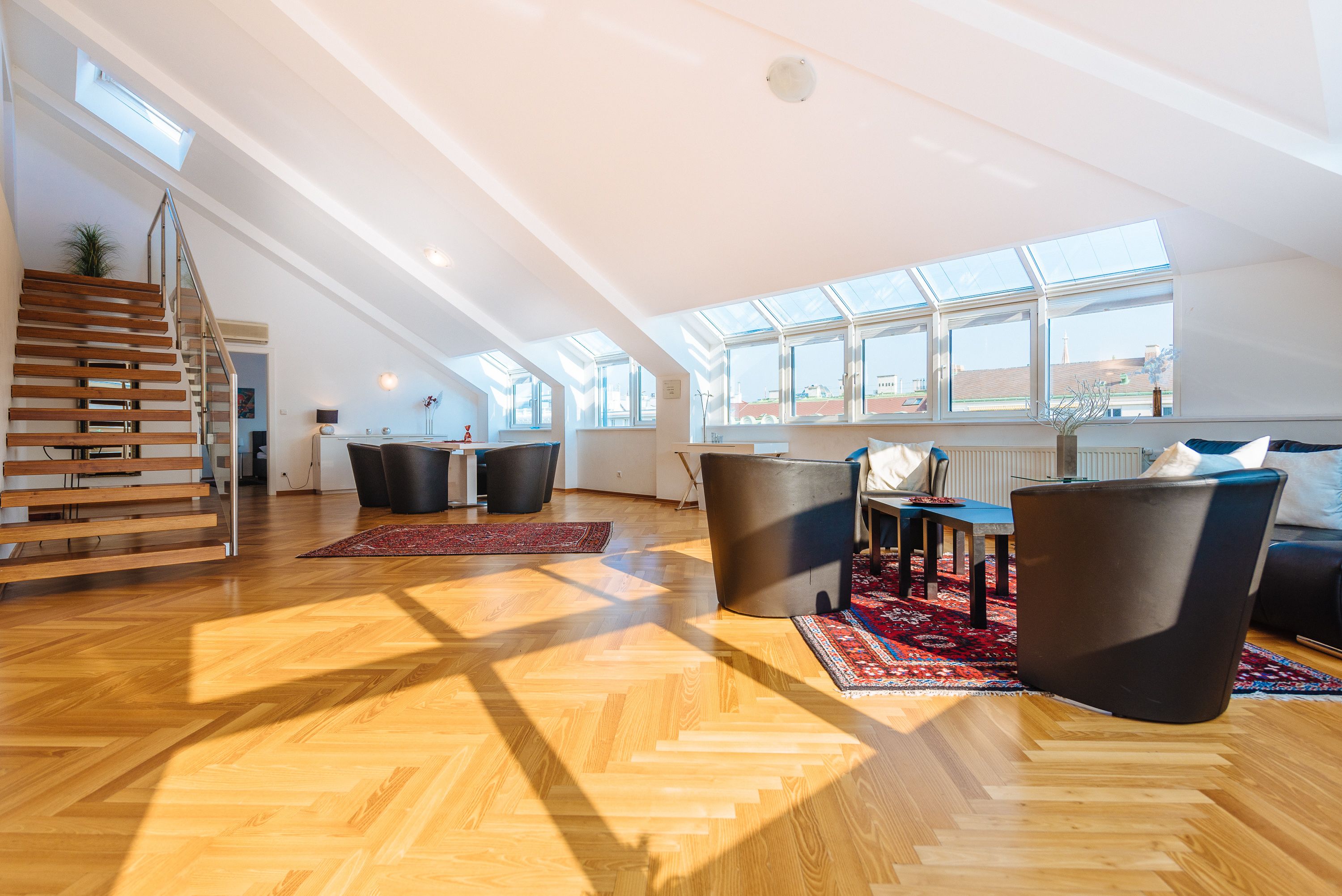 A spacious loft with a wooden staircase, a dining area with a table and chairs, and a large window providing natural light.