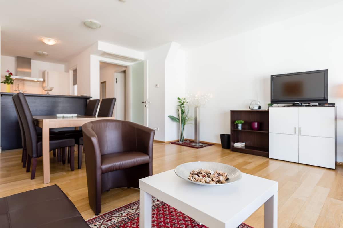 A modern living room with a dining area, a flat-screen TV, and a kitchen in the background.