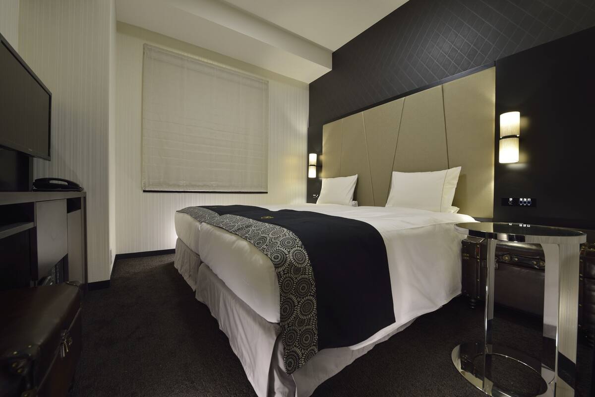 Une chambre d’hôtel avec un grand lit, une table de chevet, une télévision et une fenêtre avec des stores.