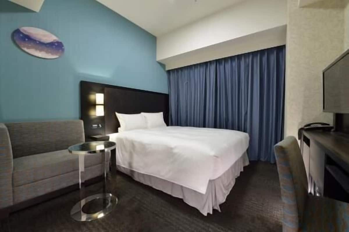 Une chambre d’hôtel avec un lit, un canapé, une table de chevet, une télévision et une grande fenêtre avec des rideaux.