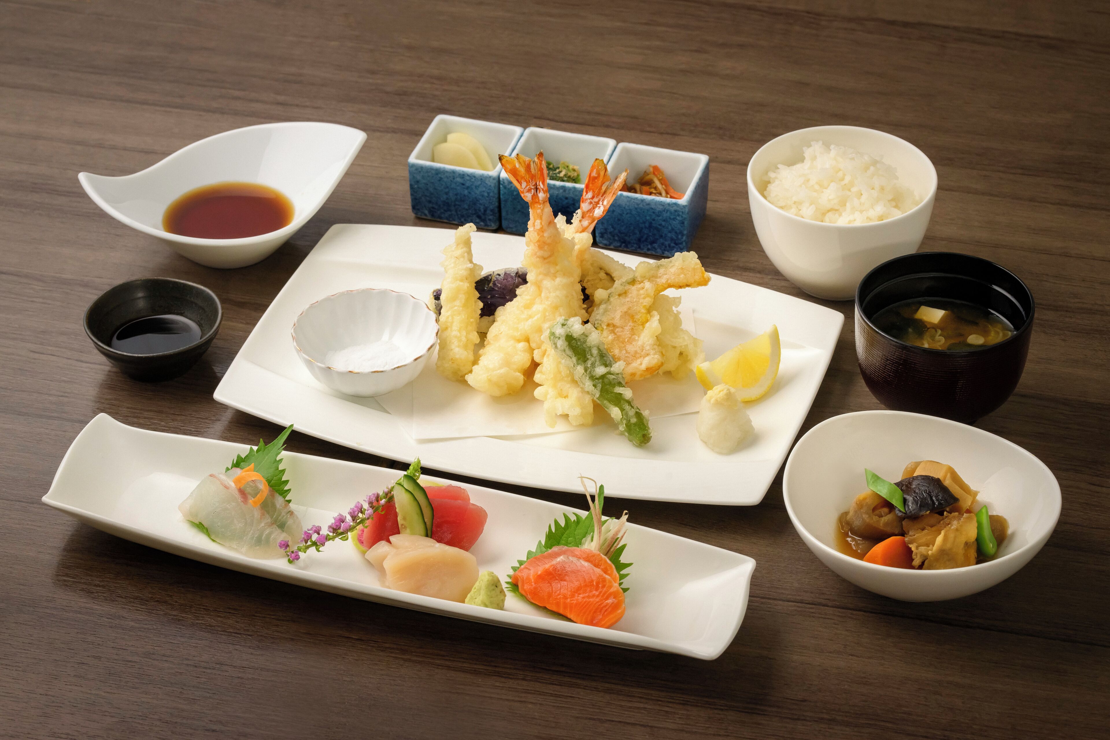 Une variété de plats japonais, notamment des beignets de tempura, des sashimis et des bols de riz, présentés sur une table en bois.