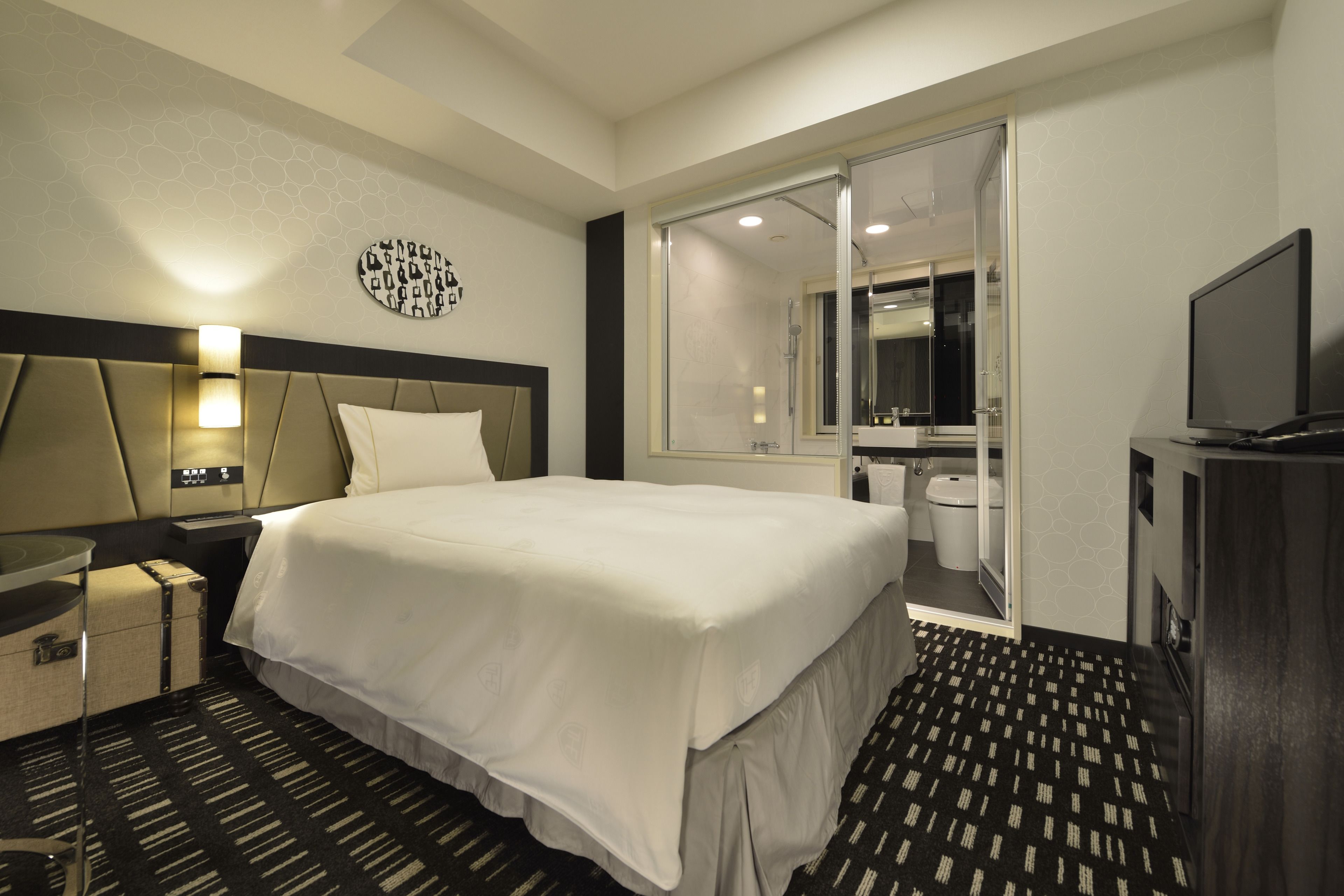 A hotel room with a large bed, a bedside table, a TV, a bathroom with a toilet and sink, and a mirror.