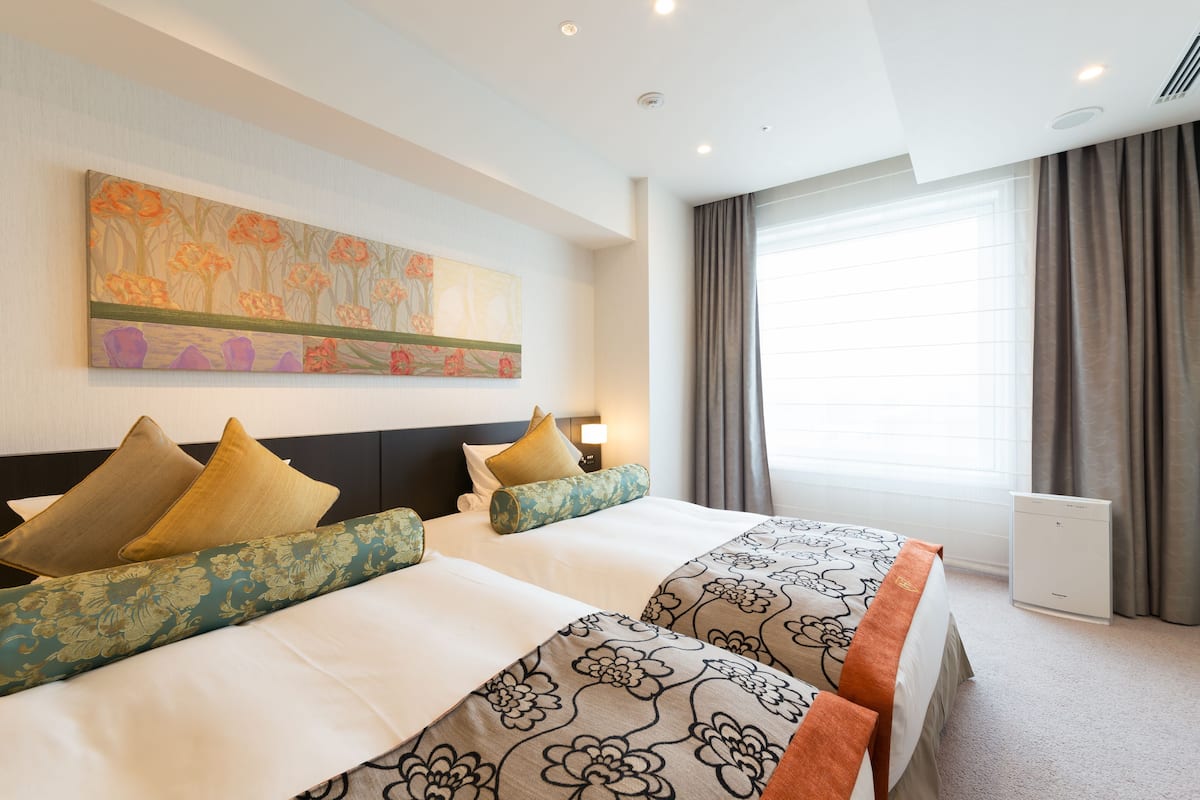Une chambre d’hôtel avec deux lits, un grand tableau floral accroché au mur, une fenêtre avec des rideaux et une table de chevet avec une lampe.