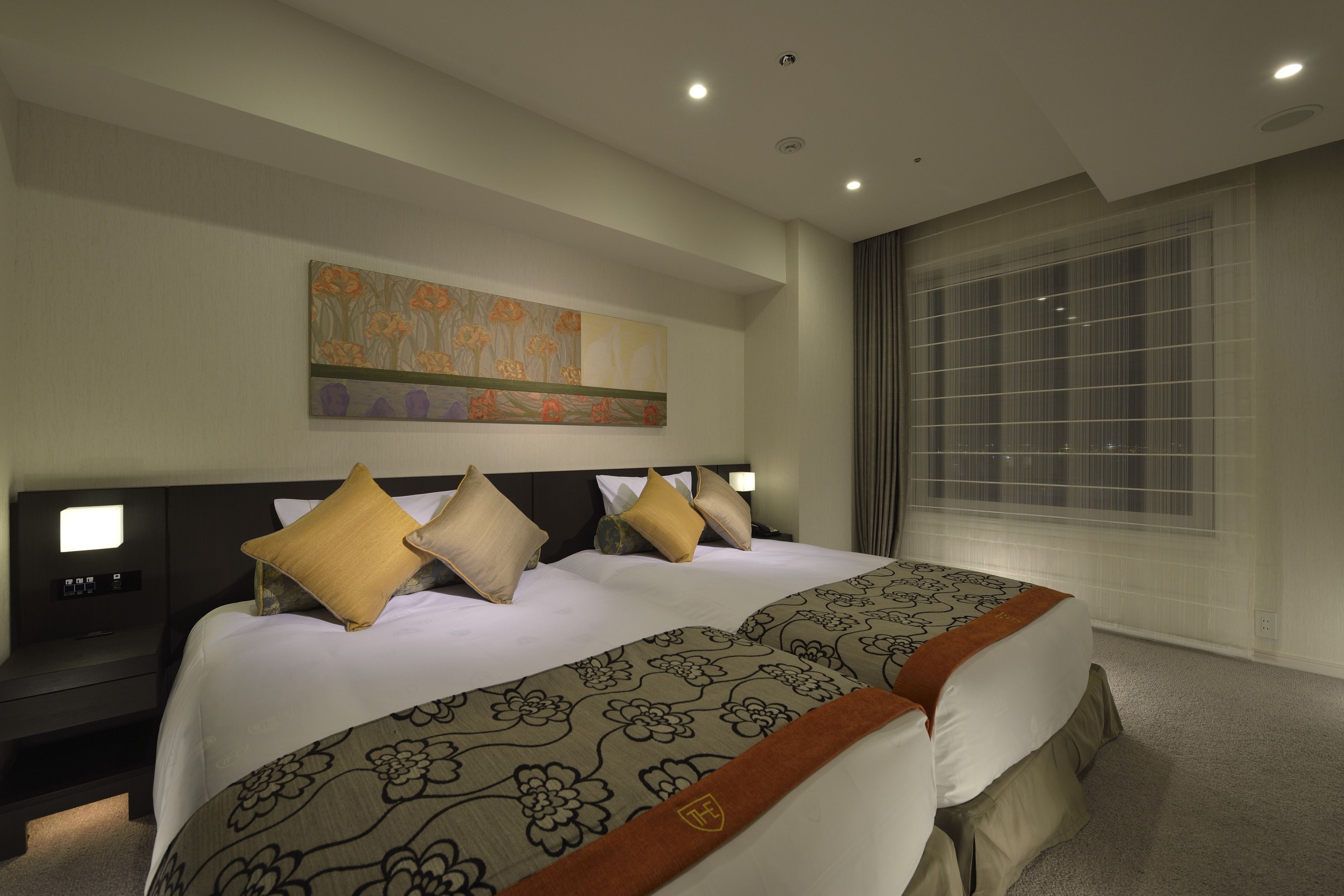 A hotel room with a large bed, a nightstand, a wall-mounted TV, and a window with blinds.