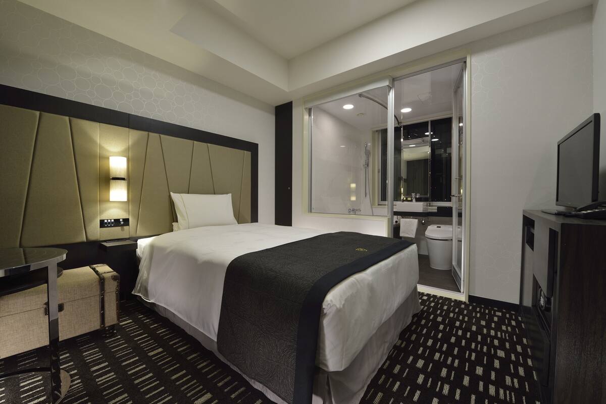 Une chambre d’hôtel moderne équipée d’un grand lit, d’un bureau, d’une chaise, d’une télévision, d’une salle de bain avec des toilettes et un lavabo, et d’un miroir.