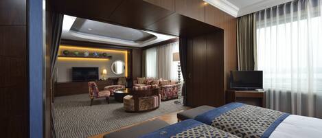 Habitación de hotel con una cama grande, televisor, zona de estar con sofá y sillones, y vistas a la ciudad desde la ventana.