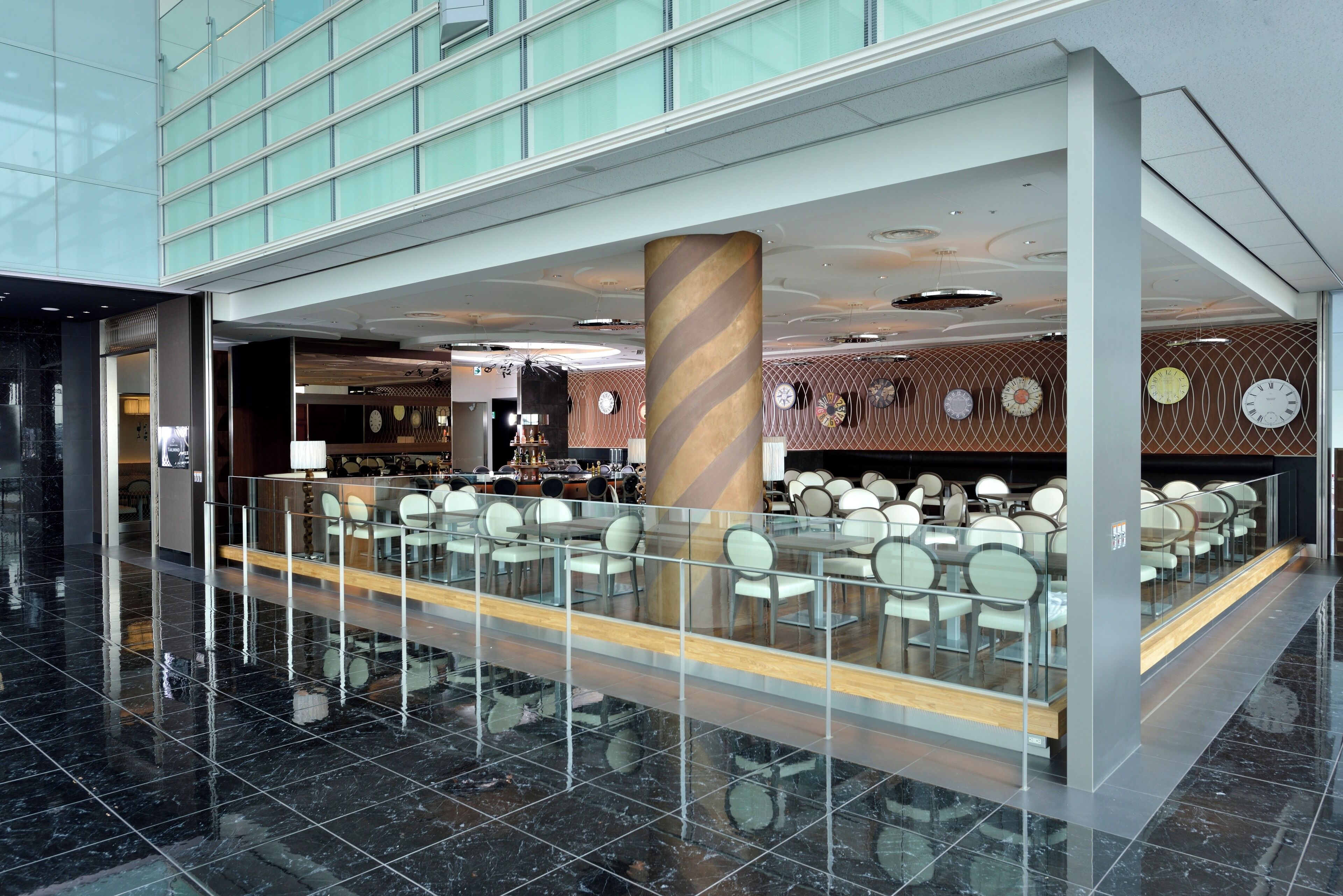 A modern hotel lobby with a glass railing, a central pillar, and a seating area with tables and chairs.
