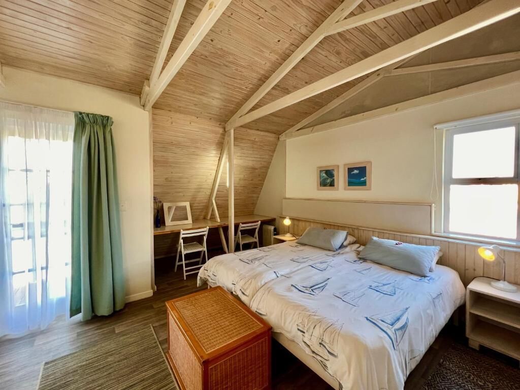 A room with a sloped ceiling, a bed with a nautical-themed bedspread, a desk with a chair, and a window with curtains.