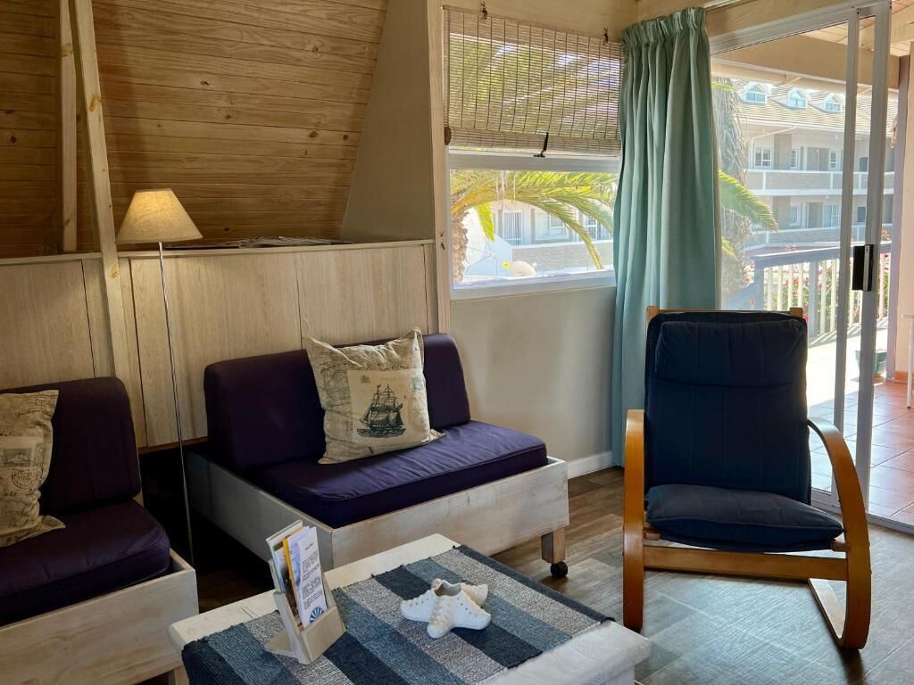 Una habitación con techo de madera, un sofá con cojines, una silla, una mesita con una revista y una ventana con vistas a edificios y palmeras.