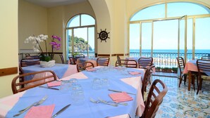 Un espace repas avec des tables disposées pour accueillir six personnes, de grandes fenêtres en arc de cercle et une vue sur la mer.