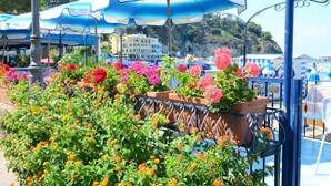 Une terrasse en bord de mer, agrémentée de parasols bleus et blancs, de plantes en pot et offrant une vue sur un bâtiment situé sur la côte.