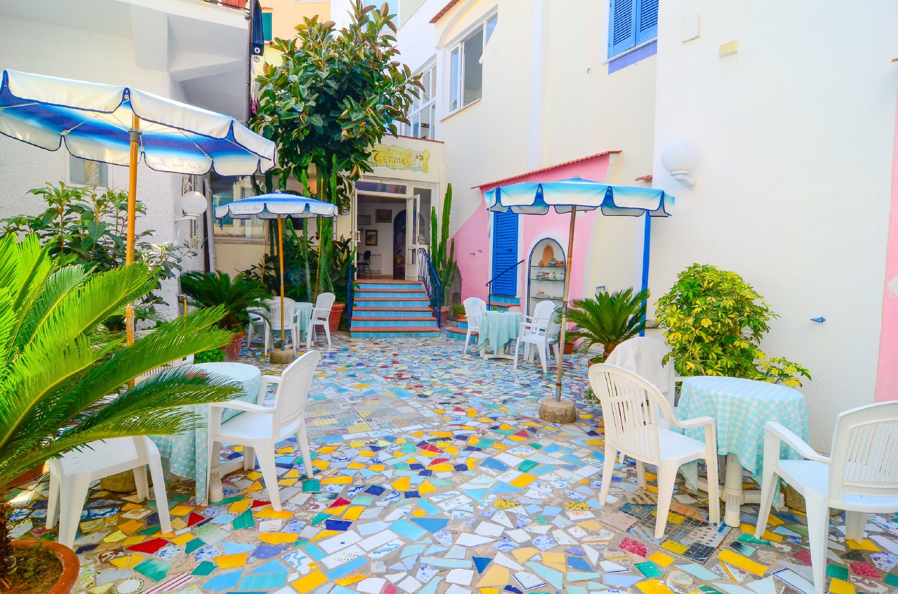 Un patio con suelo de mosaico colorido, sillas de plástico blanco y sombrillas azules.