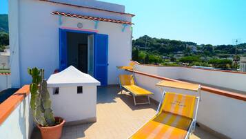 A rooftop terrace with a white building, blue shutters, a potted cactus, and two yellow-striped lounge chairs.
