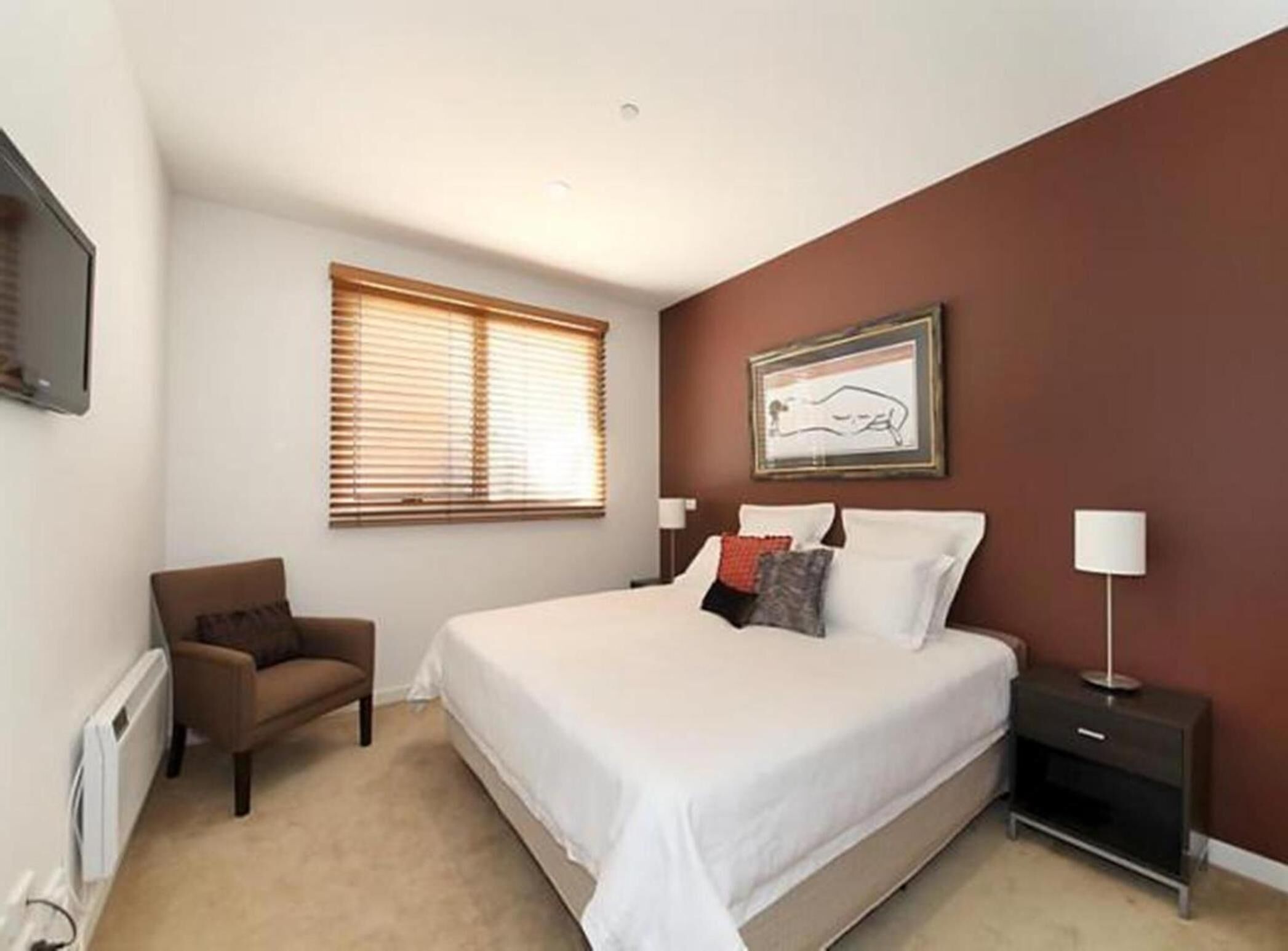 A bedroom with a large bed, a chair, a bedside table with a lamp, and a window with blinds.
