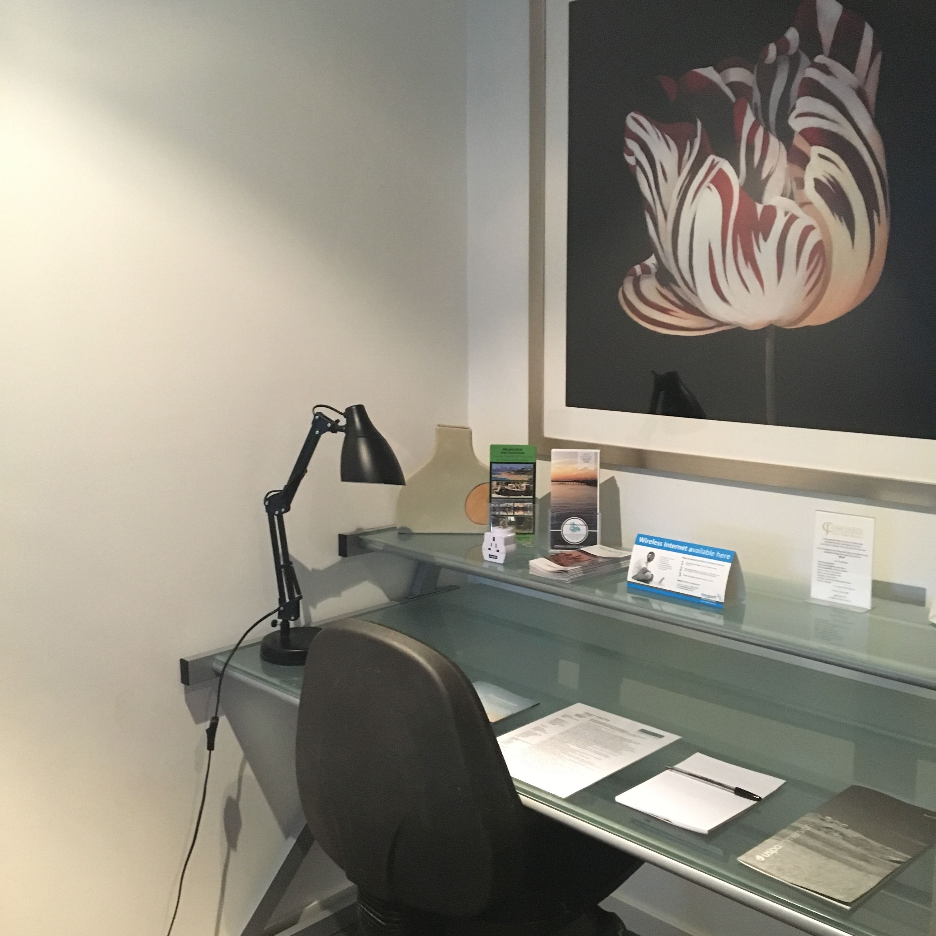 A glass desk with a black desk lamp, a chair, and various items including a framed picture, a small box, and some papers.