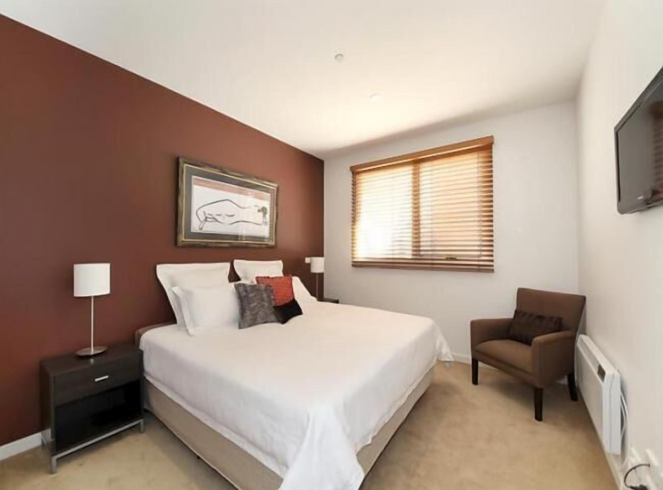 A bedroom with a bed, a nightstand, a chair, a window with blinds, and a framed picture on the wall.