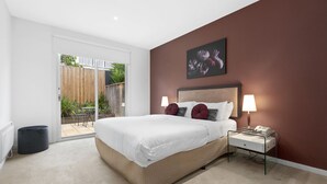 A bedroom with a large bed, a nightstand, a wall-mounted lamp, and a sliding glass door leading to an outdoor area. - Brighton Bay Apartments (Brighton)
