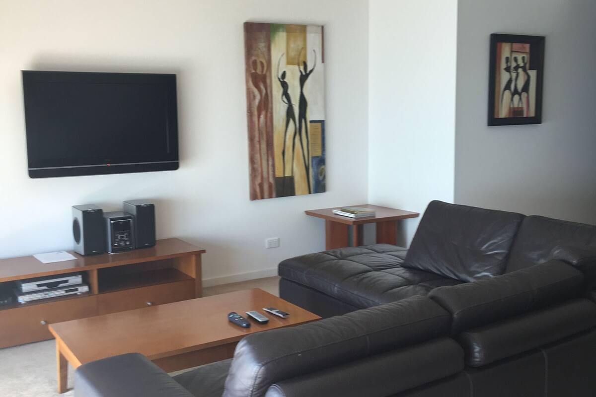 A modern living room with a black leather sofa, a wooden coffee table, a flat-screen TV mounted on the wall, and two framed abstract paintings.