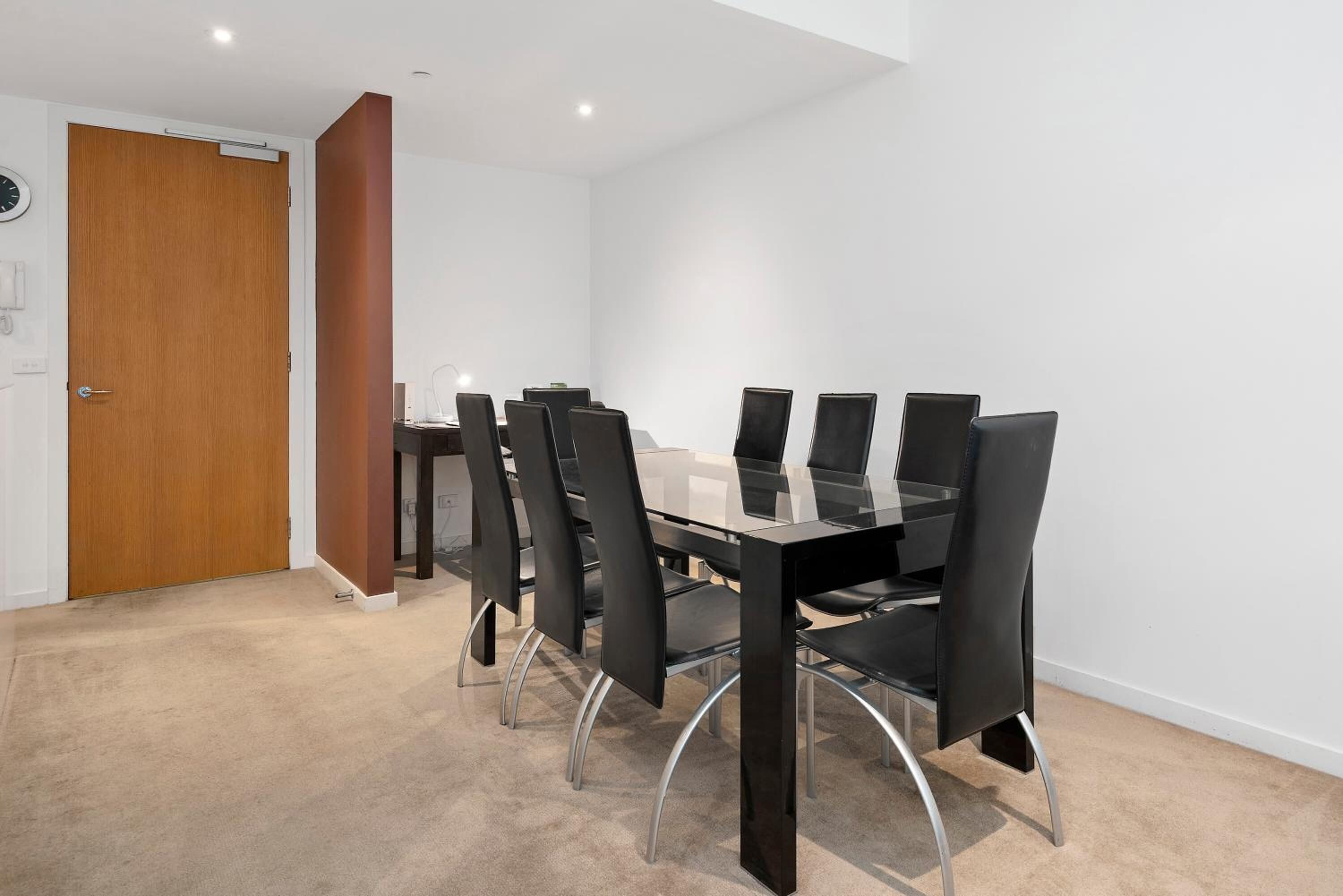 Una sala conferenze con un tavolo in vetro e sedie nere, una porta in legno e un muro bianco.