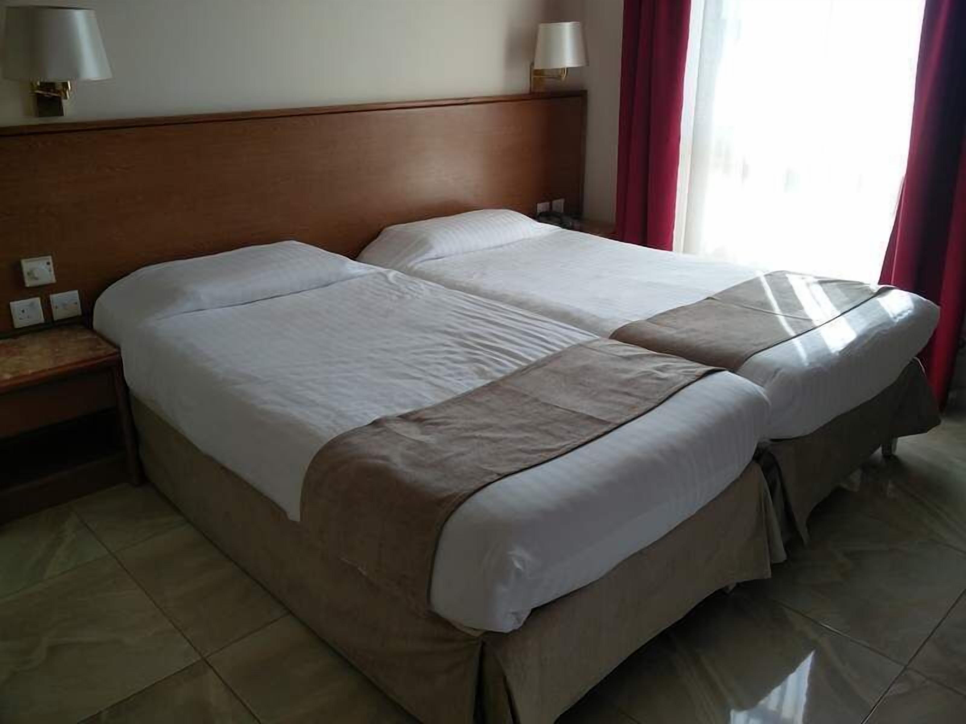 A double bed with a wooden headboard, a nightstand with a lamp, and a window with red curtains.