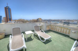A rooftop terrace with two lounge chairs and a small table, overlooking a cityscape with buildings and a clear sky.