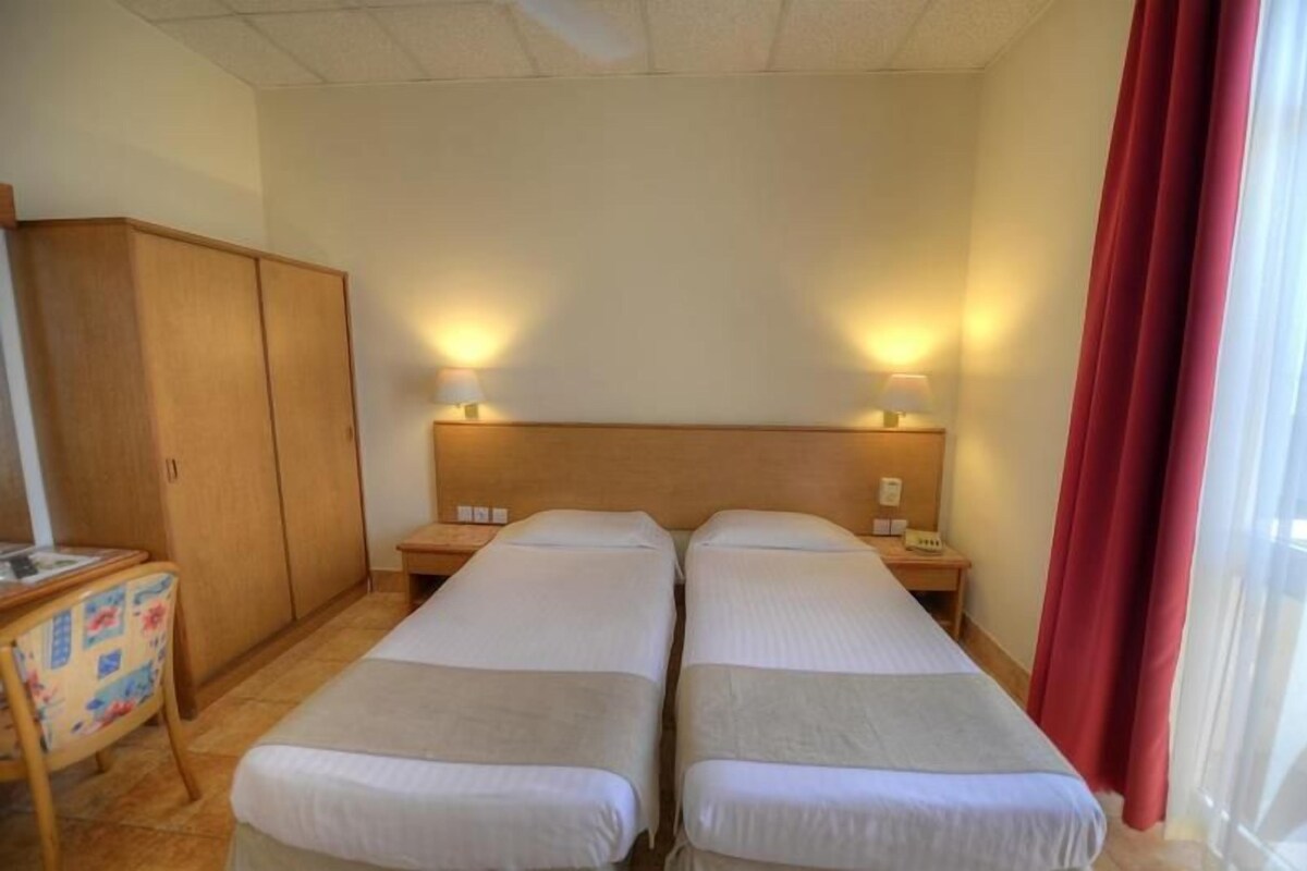 A hotel room with two single beds, a wooden wardrobe, a desk with a chair, and bedside lamps.