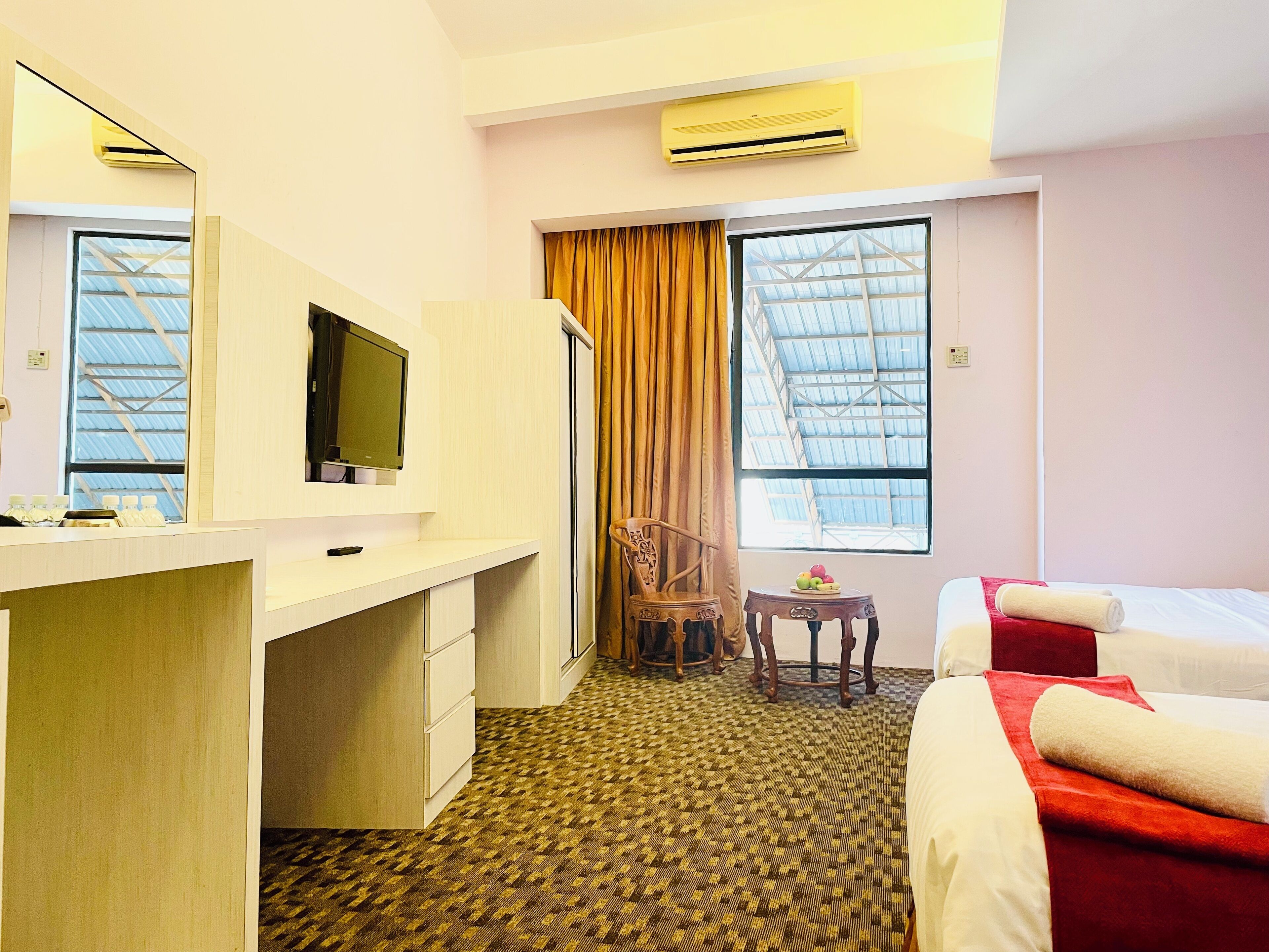 A hotel room with a bed, a desk with a flat-screen TV, a chair, a small table with a fruit bowl, and a large window with curtains.