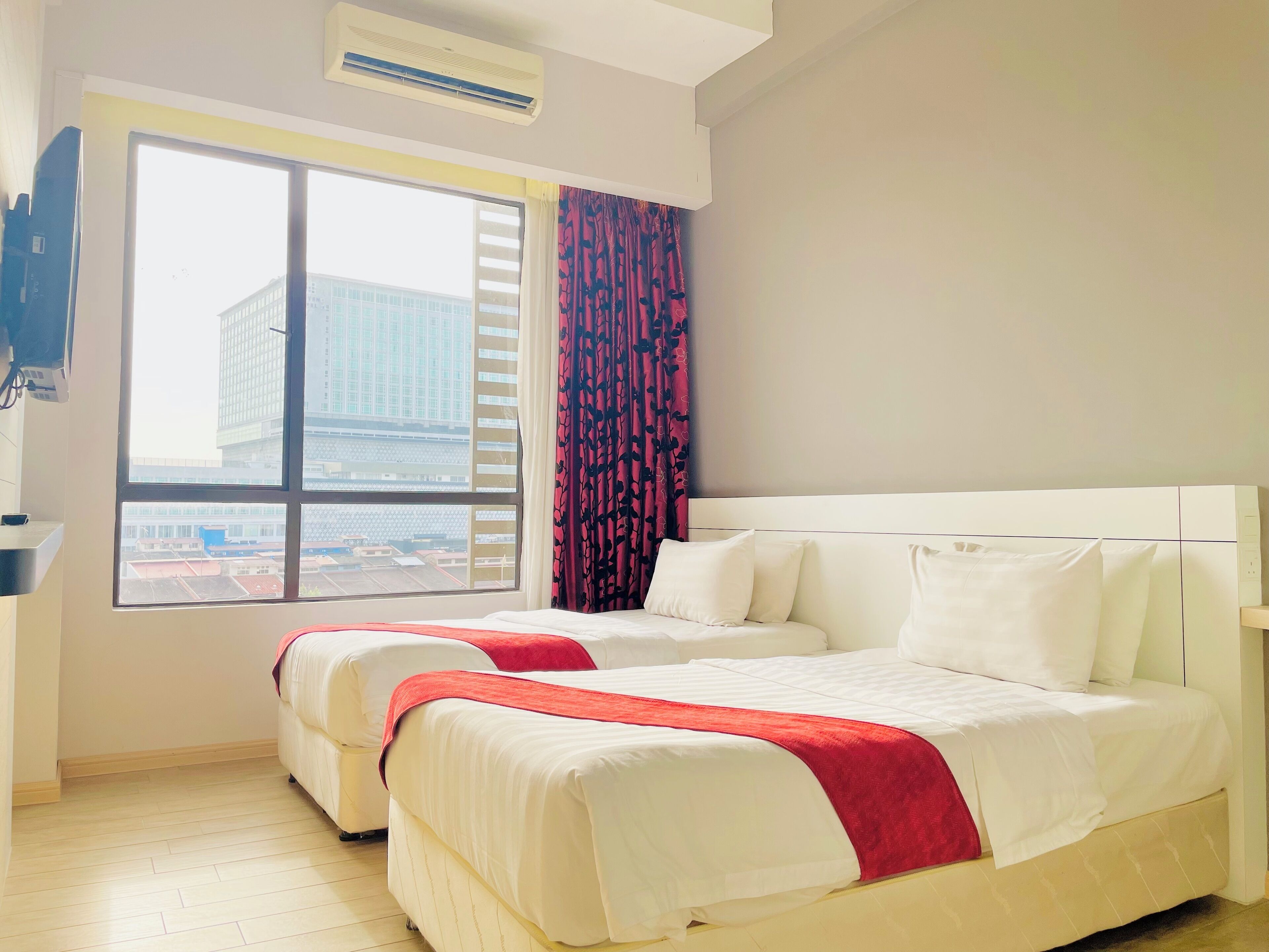 A hotel room with two beds, a large window with blinds, a red curtain, and a view of buildings outside.