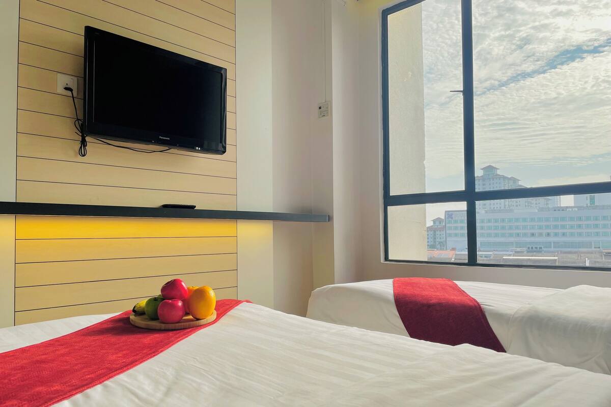A hotel room with two beds, a flat-screen TV mounted on the wall, a fruit basket on the nightstand, and a large window offering a city view.