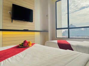 Une chambre d’hôtel avec deux lits, un téléviseur à écran plat fixé au mur, un panier de fruits sur la table de chevet et une grande fenêtre donnant sur la ville.
