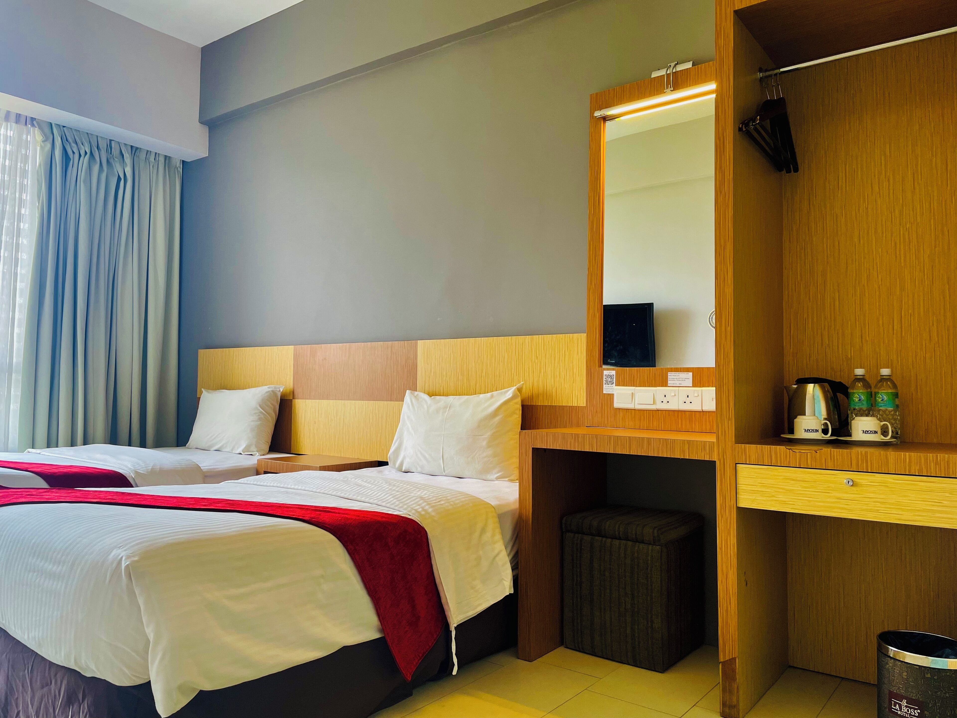 A hotel room with two beds, a wooden headboard, a mirror, a small table, a trash bin, and a shelf with toiletries.