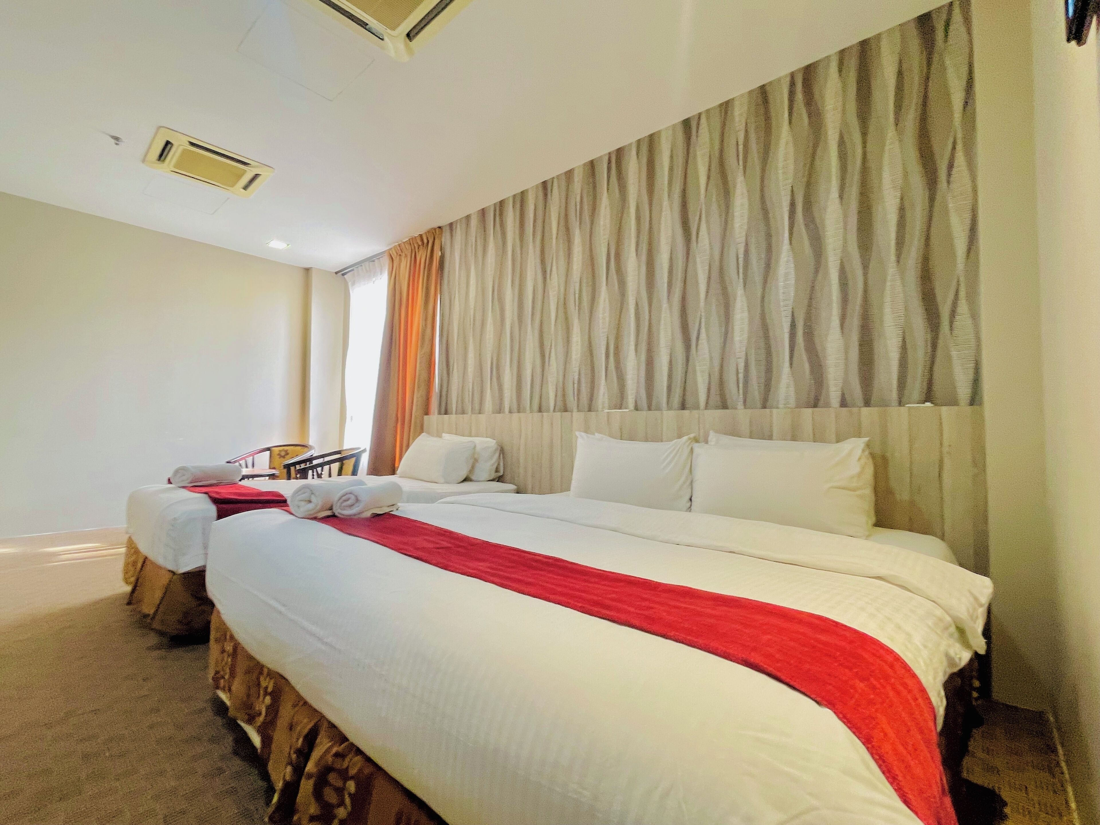 A hotel room with two beds, a large window with curtains, a wooden headboard, and a red towel on one bed.