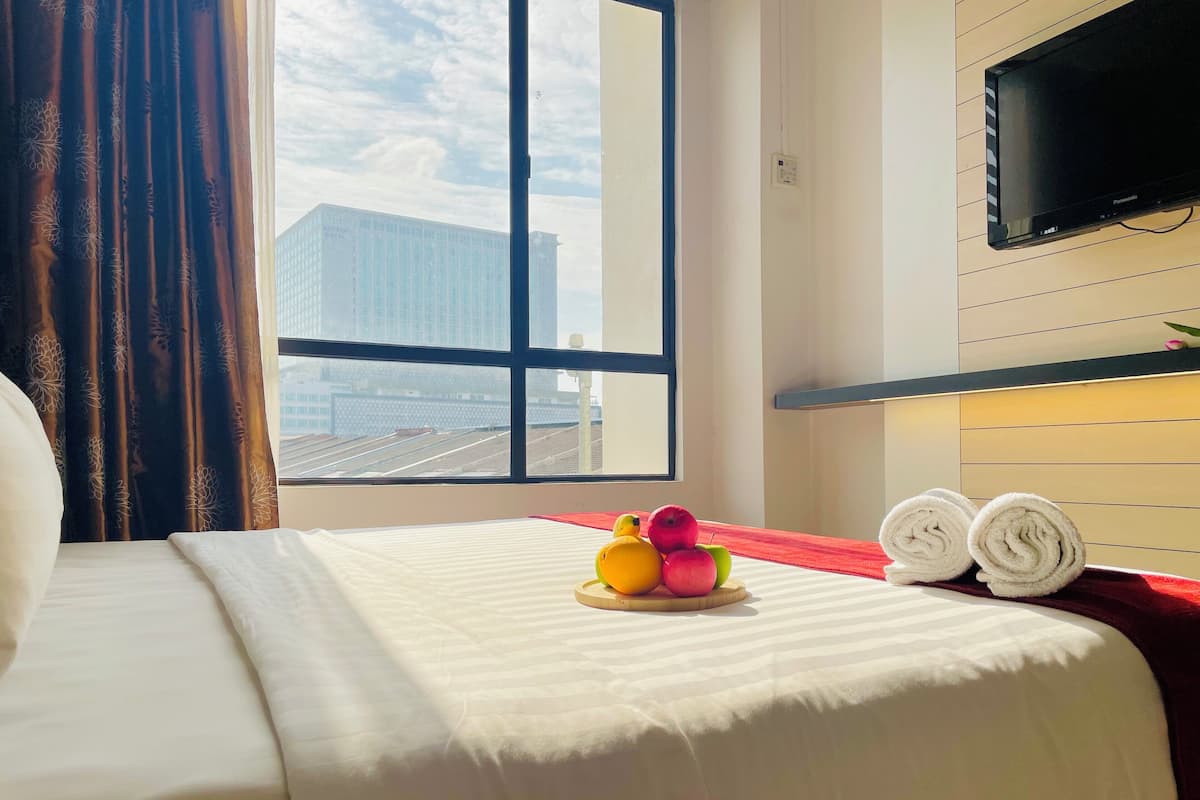 A hotel room with a bed, a window with a city view, a flat-screen TV mounted on the wall, and a small table with fruit and towels.