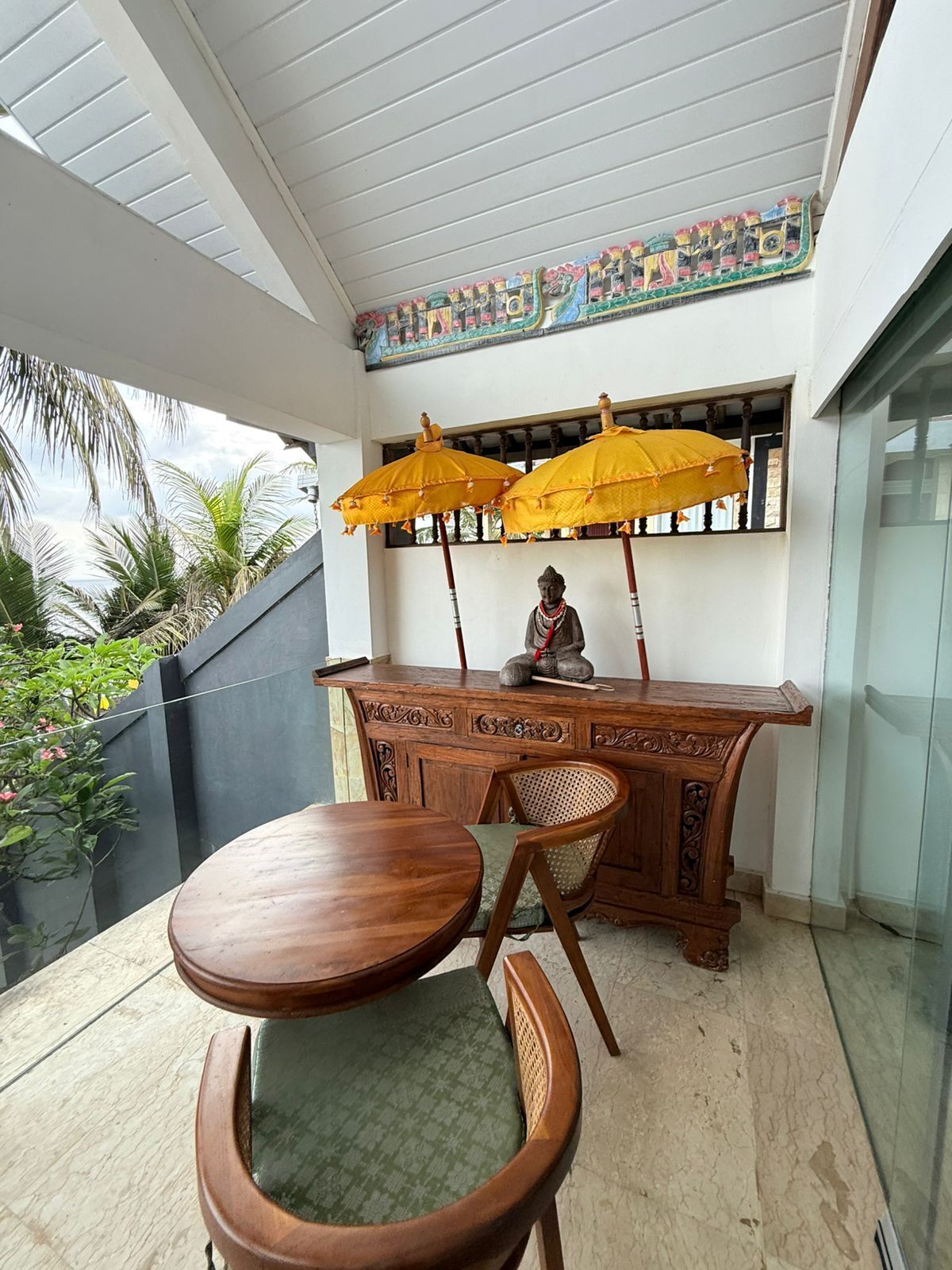 A wooden table and chair set on a balcony with a decorative statue and two yellow umbrellas.
